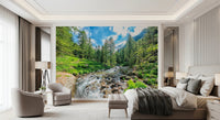 Alpine Stream wallpaper mural with a forest stream behind a taupe bed and beige armchairs in a bedroom
