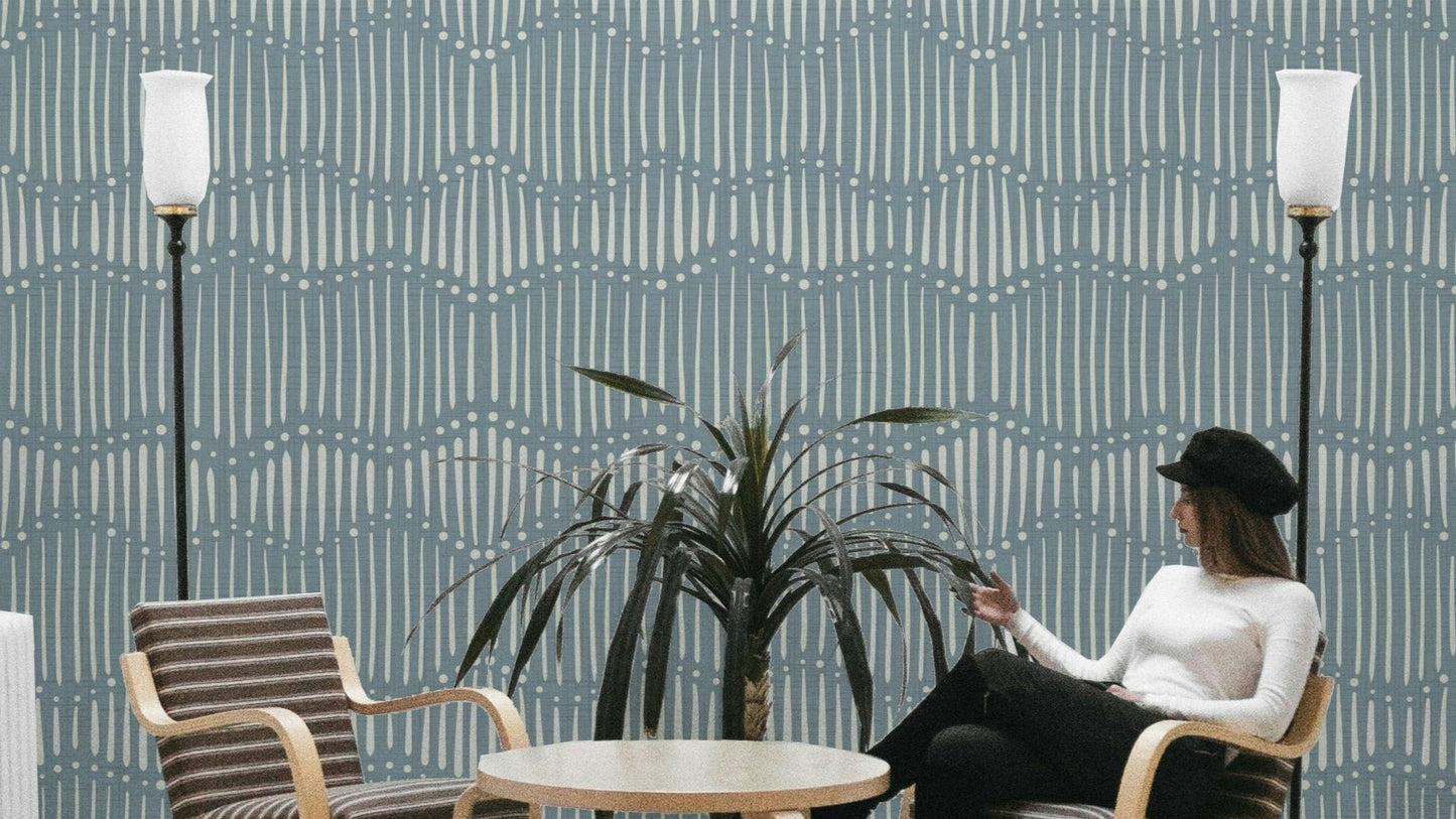 Boho Waves On Arona Canvas wallpaper in slate blue with cream dash lines behind a woman seated in a lounge with floor lamps