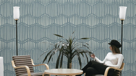 Boho Waves On Arona Canvas wallpaper in slate blue with cream dash lines behind a woman seated in a lounge with floor lamps
