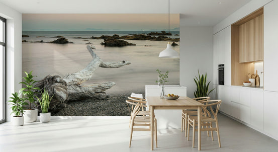 Shoreline Symphony wallpaper mural of dusk driftwood on a pebble beach behind a wood dining table in a kitchen