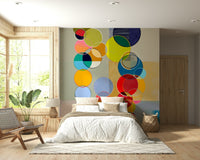 Chromatic Circles wallpaper mural of bright circles behind a white bed and woven lounge chair in a bedroom