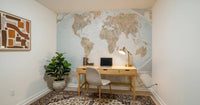 Classic world map in blue and earthy tones wallpaper mural behind a wood desk with a laptop and brass lamp in a home office