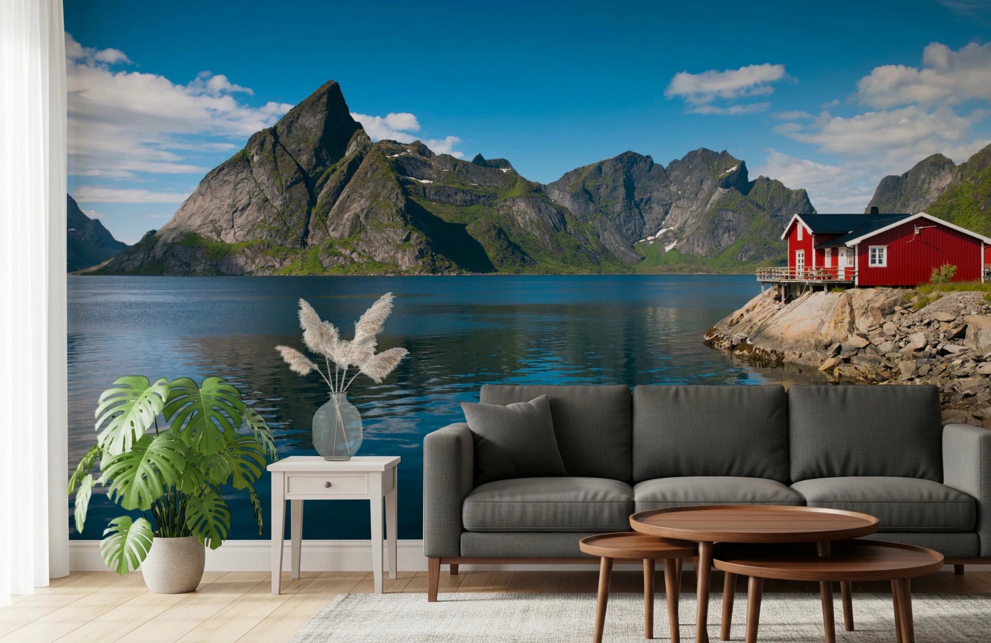 Fishing huts wallpaper mural of grey mountains and a red cabin behind a grey sofa and monstera plant in a living room