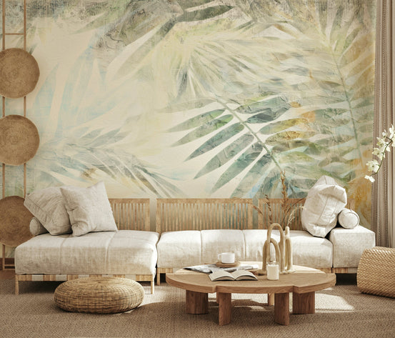 Gilded Fronds wallpaper mural of soft sage palm leaves behind a low cream wooden-frame sofa with rattan wall discs in a living room