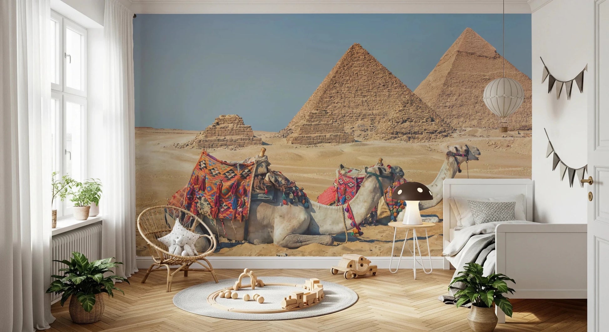 Giza wall mural of the pyramids and saddled camels in a nursery with a white bed, rattan chair and wood toys