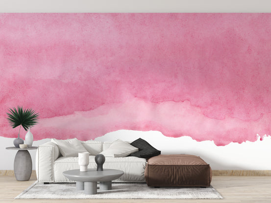Pink shades watercolor wallpaper with a soft pink wash behind a white sofa and brown leather ottoman in a living room