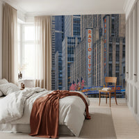 Radio City's Glow wallpaper mural of the Radio City sign and NYC towers behind a white bed with rust throw in a bedroom