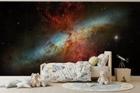 Stardust Symphony wallpaper mural of a fiery starburst galaxy in a kids room with rocket bedding and plush toys