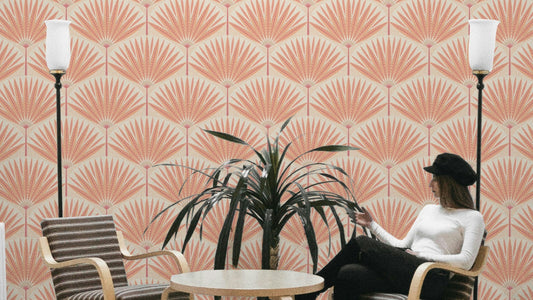 Sunset Palm Paradise wallpaper in terracotta palm fans behind a woman in a wood armchair with a dracaena in a living room