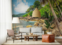 Tropical Shores wallpaper mural with palms and blue sea behind a grey sofa and leather pouf in a living room