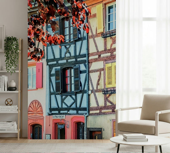 Vintage Alsace wallpaper mural of colorful half-timbered houses beside a beige armchair and bookshelf in a living room